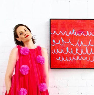 Australian artist Nadia Düsselberg stands in front of one of her paintings. Nadia wears a bright red dress with pink dots, which matches the bright red of the painting behind her. The painting is red with blue scribbles.