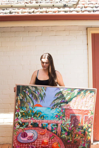 Stephanie Perrins dressing in black,  holding a colourful piece of artwork, standing under the shades.