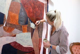 Artist Krystal Bradford hold a paintbrush in one hand and a jar of paint in the other as she works on a large abstract artwork in reds, oranges, charcoal greys and pale beige.