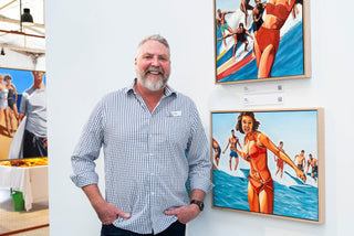 Artist Pete James wears a blue and white check shirt and stands in front of two of his artworks. Each work is a painting of a group of people surfing a wave.