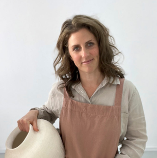 Sculpture artist Cara Kushlin wears a muted pink apron and light colour shirt. She stands holding a large piece of ceramic pottery.