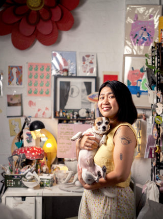 Sculpture artist Yang Qiu stands in her studio. She wears a yellow top and holds a small dog.