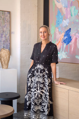Art to Art Founder Philippa Demase wearing a black and white dress and standing in front of original artworks hanging in the Art to Art gallery.